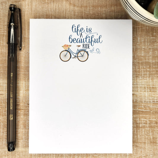 Life is a beautiful ride words with a cute bike with flowers. Notepad on wooden background with black pen next to it.