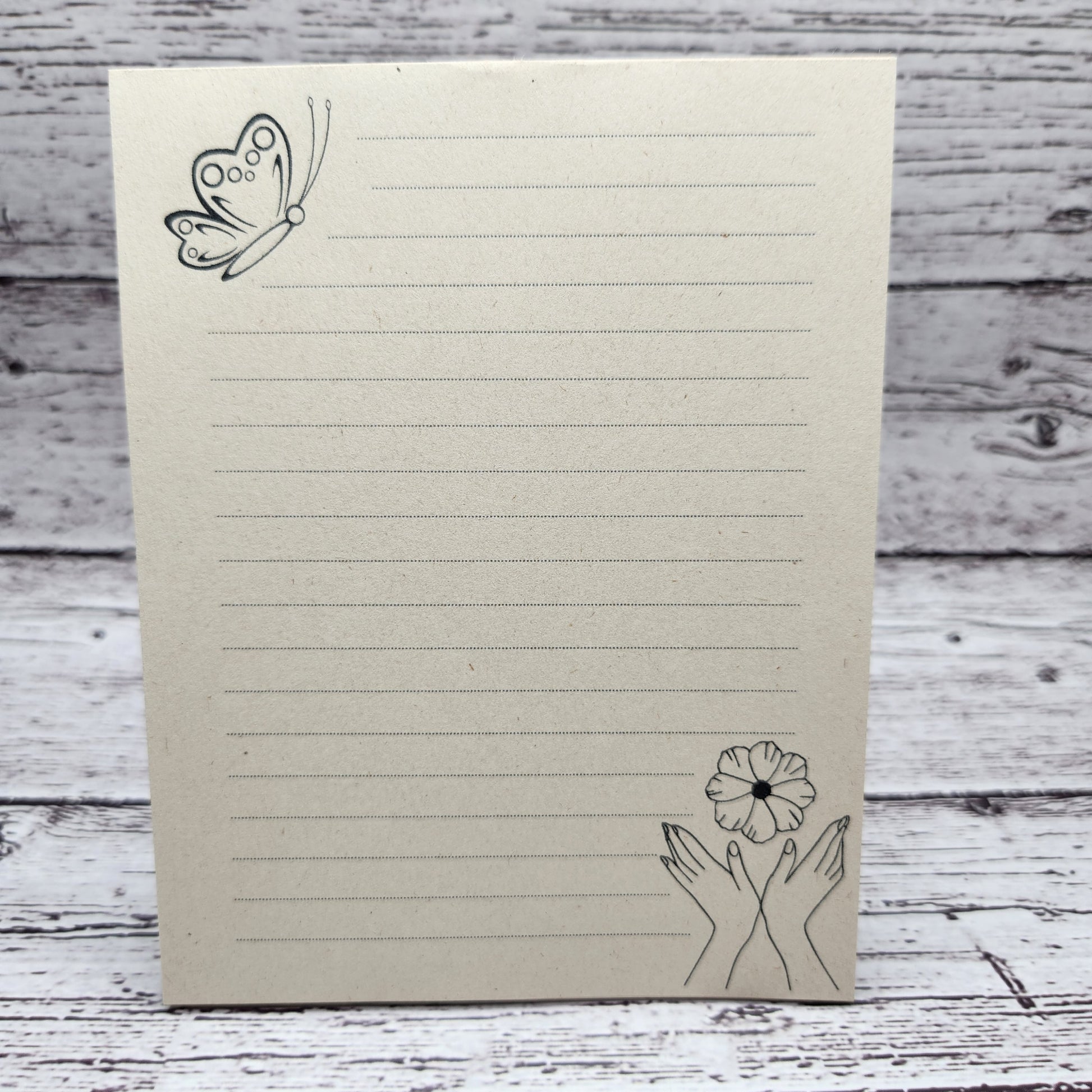 Soft beige sugarcane paper notepad on white barn board background.  Note pad is lined and has a butterfly accent on the upper left corner and hands with flowers on the bottom right corner.  all designs are line drawings.