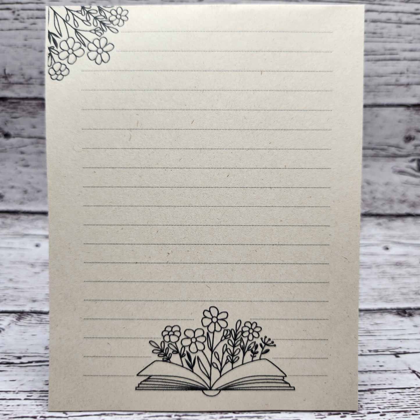Soft beige sugarcane paper notepad on white barn board background.  Note pad is lined and has flower accent on the upper left corner and open book with flowers coming out of the center on the bottom center of the page. all designs are line drawings.