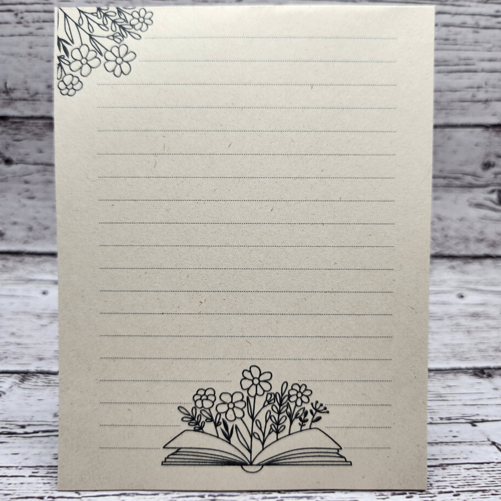 Soft beige sugarcane paper notepad on white barn board background.  Note pad is lined and has flower accent on the upper left corner and open book with flowers coming out of the center on the bottom center of the page. all designs are line drawings.