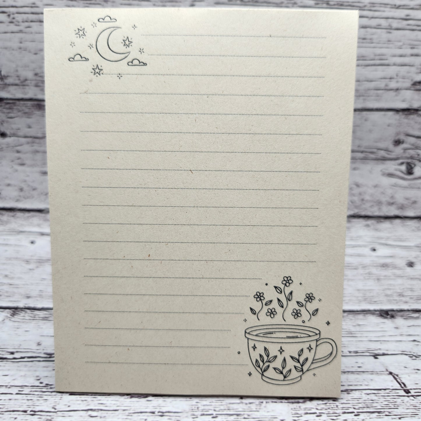 Soft beige sugarcane paper notepad on white barn board background.  Note pad is lined and has moon accent on the upper left corner and cup with flowers on the bottom right corner.  all designs are line drawings.