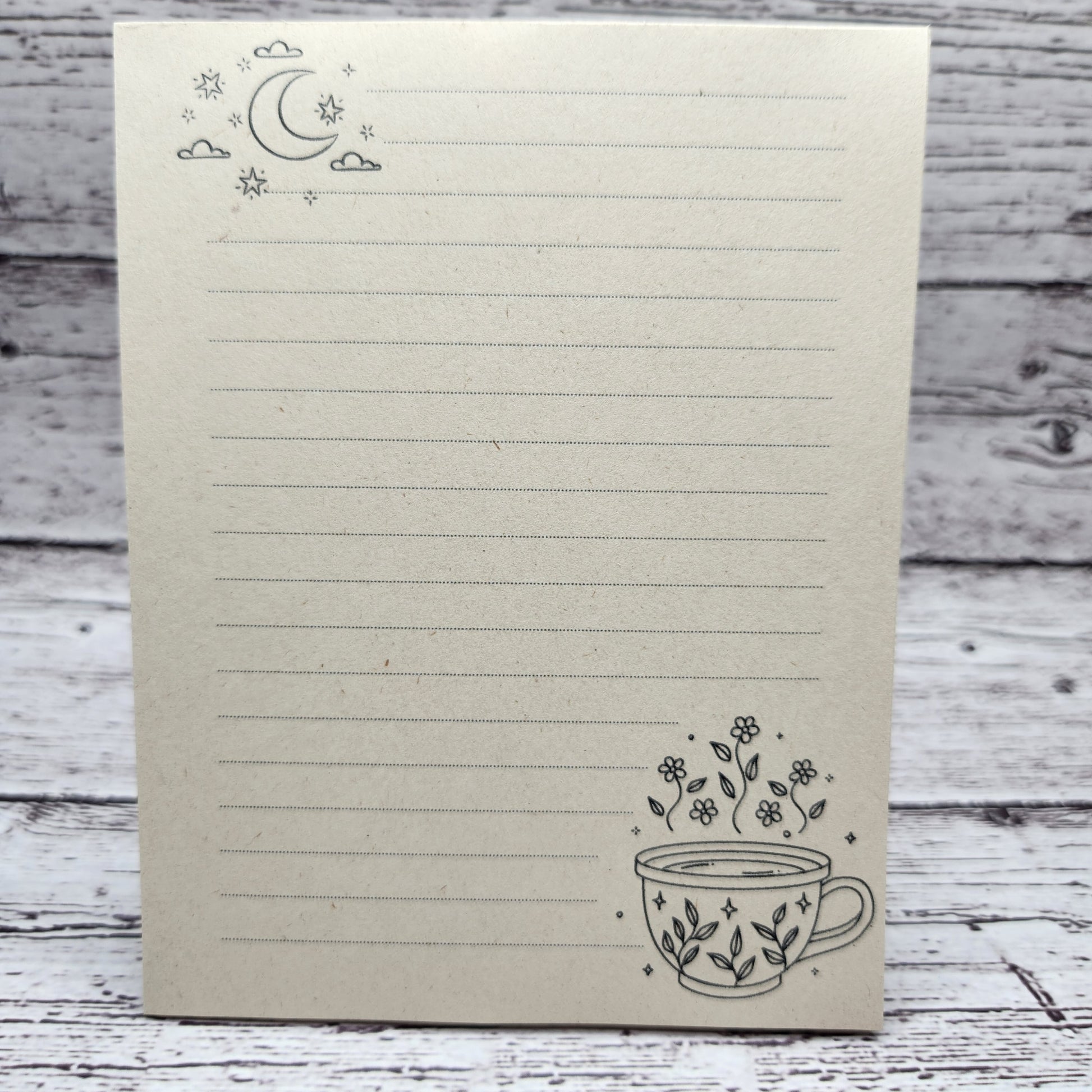 Soft beige sugarcane paper notepad on white barn board background.  Note pad is lined and has moon accent on the upper left corner and cup with flowers on the bottom right corner.  all designs are line drawings.