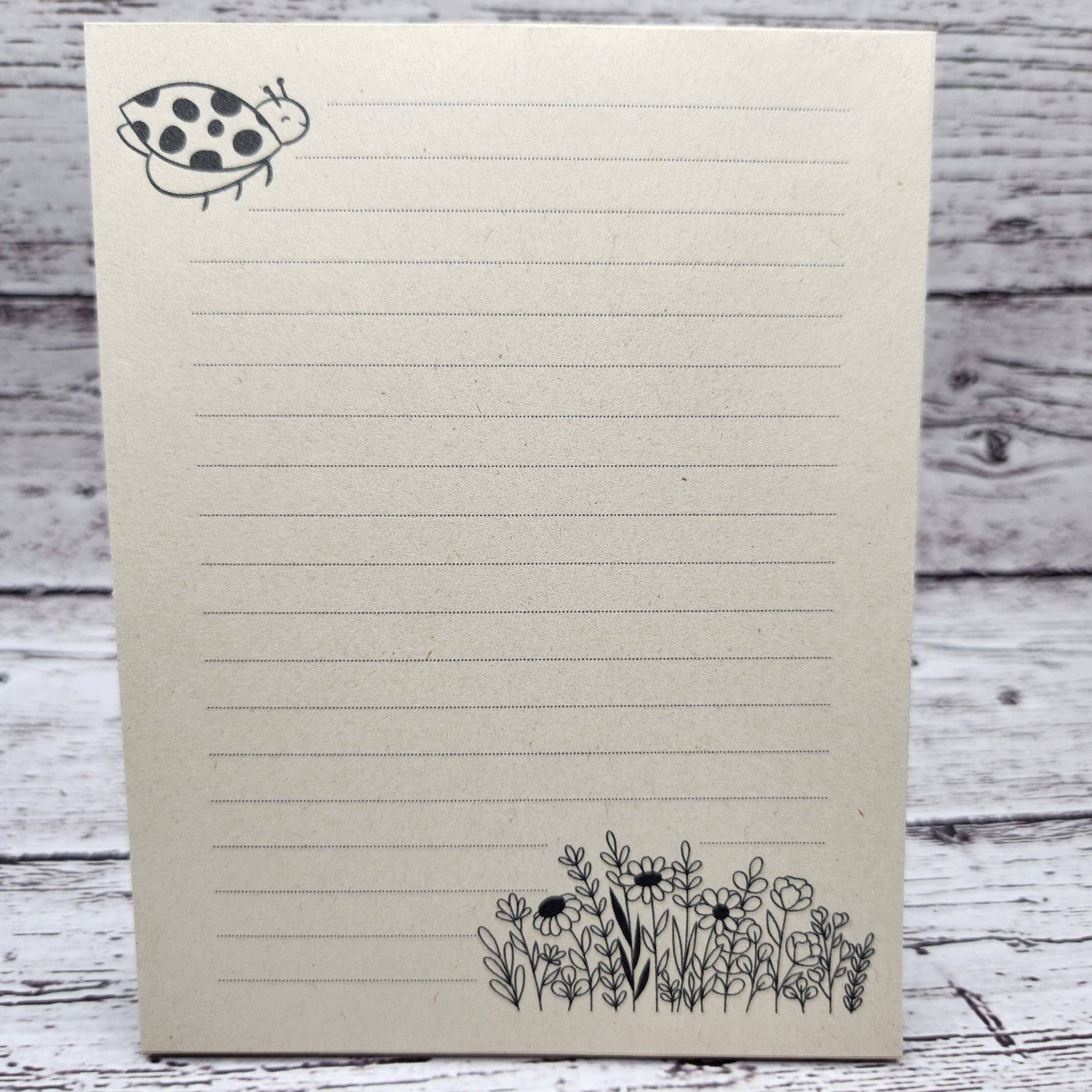 Soft beige sugarcane paper notepad on white barn board background.  Note pad is lined and has ladybug accent on the upper left and flowers on the bottom right corner.  all designs are line drawings.