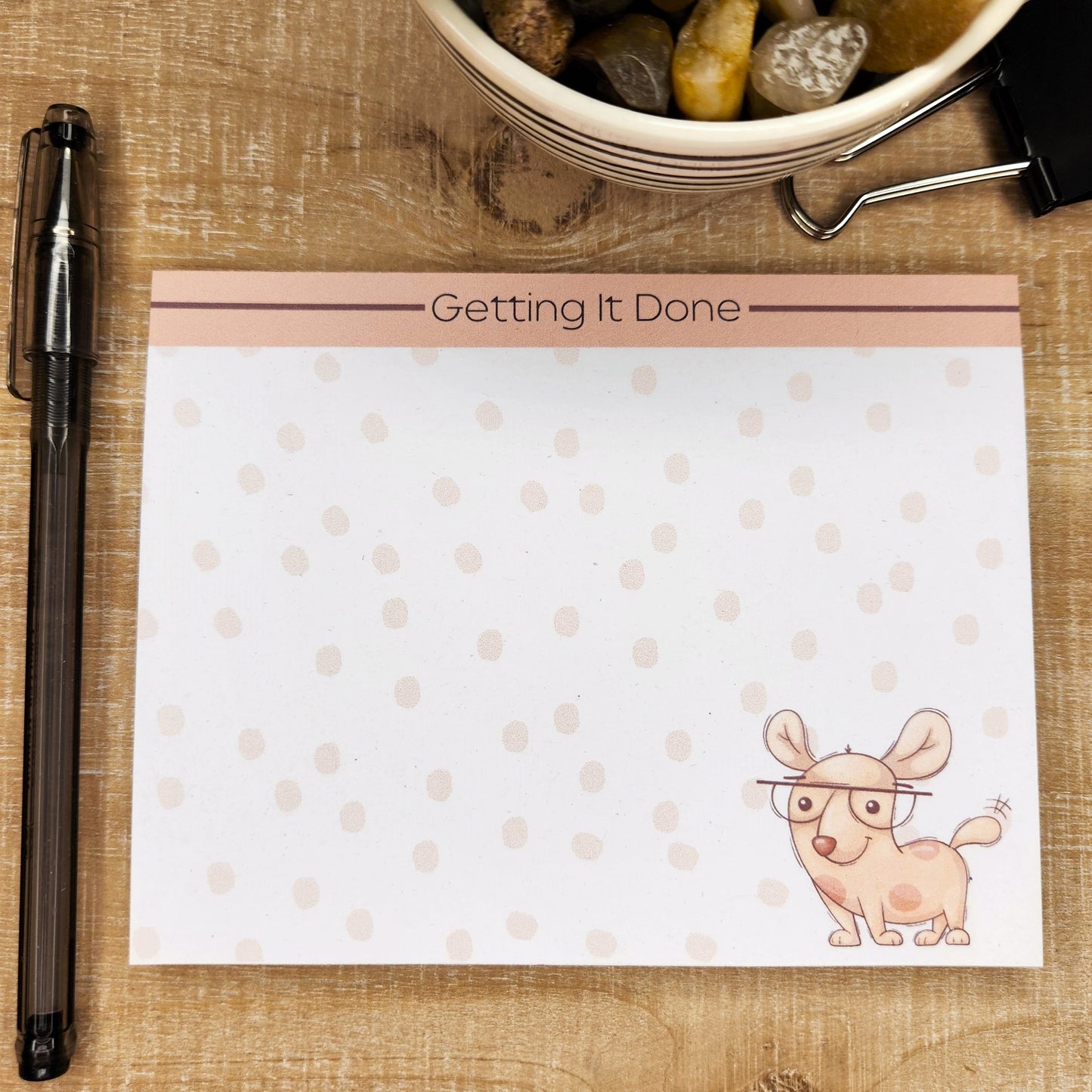 White notepad on wood background. Top of page has blush accent bar with burgundy line. The words on the center top of the page are ""Getting It Done". Standing puppy with glasses is on the bottom right side of the page. Faint dots are though out the page.