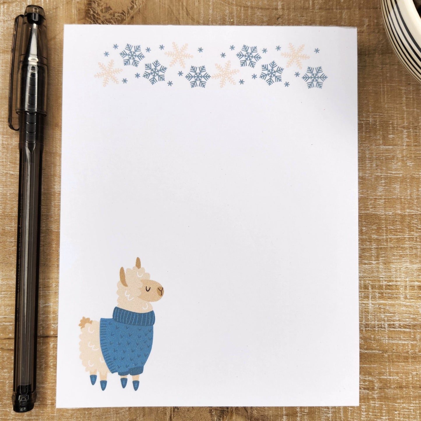 Fluffy Alpaca wearing a blue sweater and blue socks in on the bottom left corner and blue and tan snowflakes line the top of the notepad.