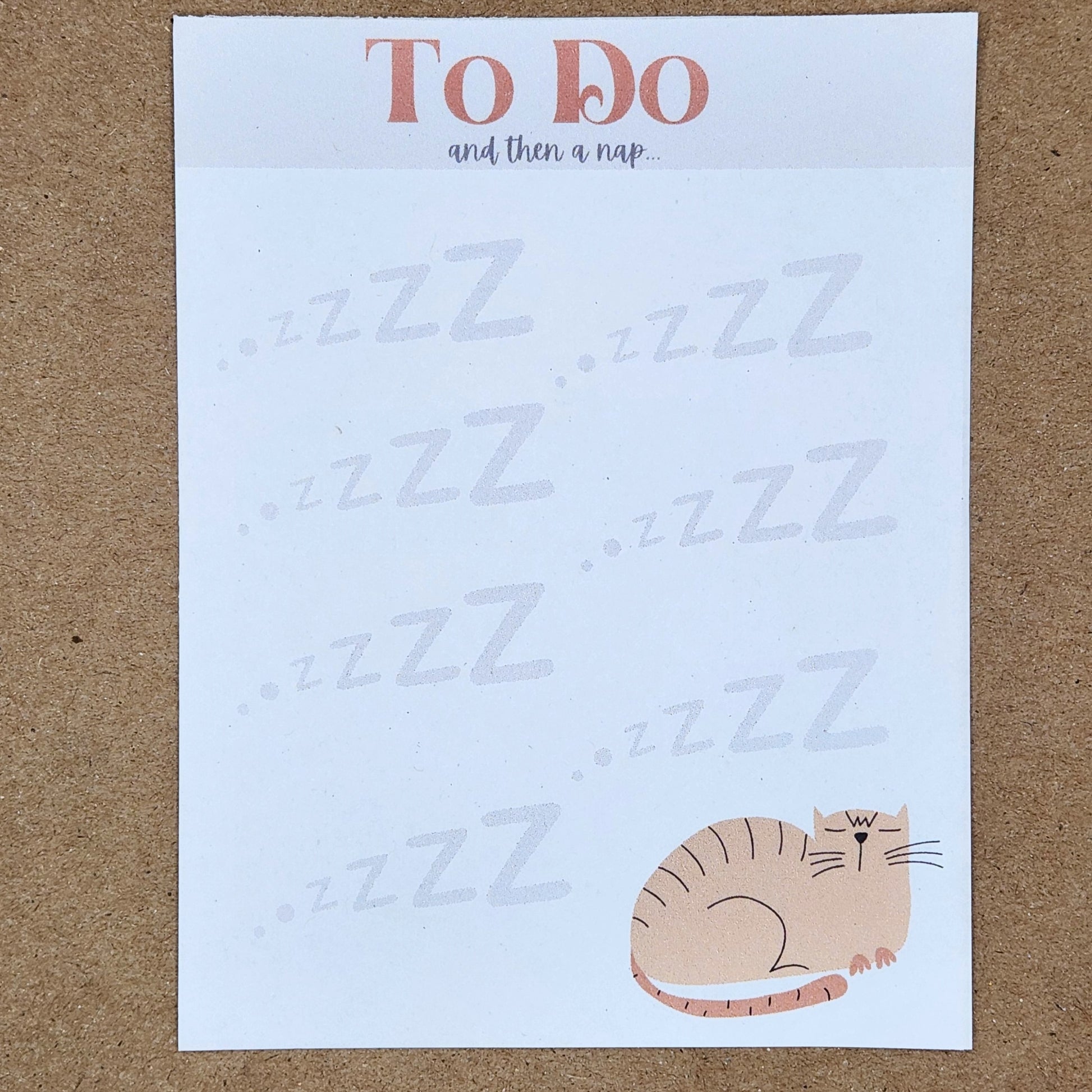 sleeping cartoon kitty is on the bottom right side of the page with zzZs all over the page.  At the top of the the page is a large To Do with the small words and then a nap.. under it. White notepad is pictured on a tan background.