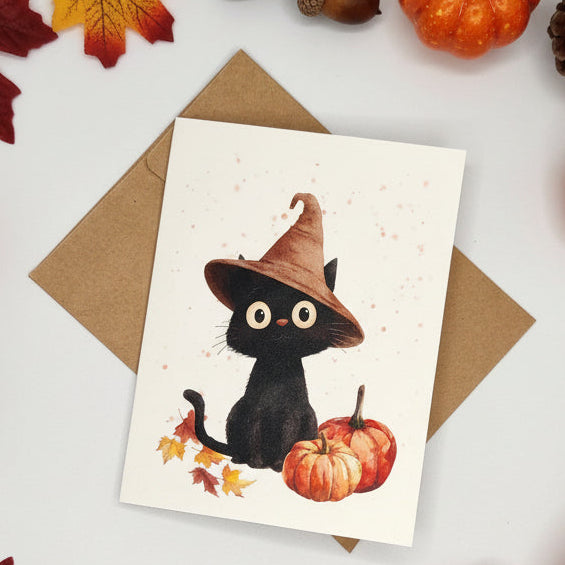 A white greeting card featuring a cute black kitten wearing a brown witch hat, sitting beside two orange pumpkins and autumn leaves, with a brown envelope behind it on a light gray background.