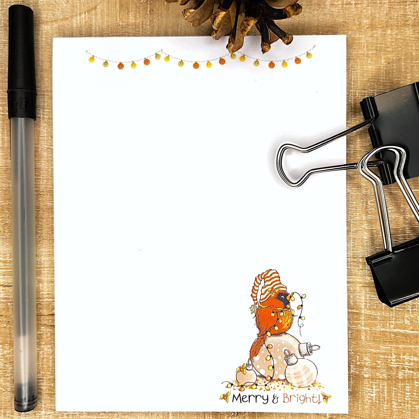 Notepad with a snowman illustration and 'Merry & Bright' text, pen, and clips on a wooden surface.