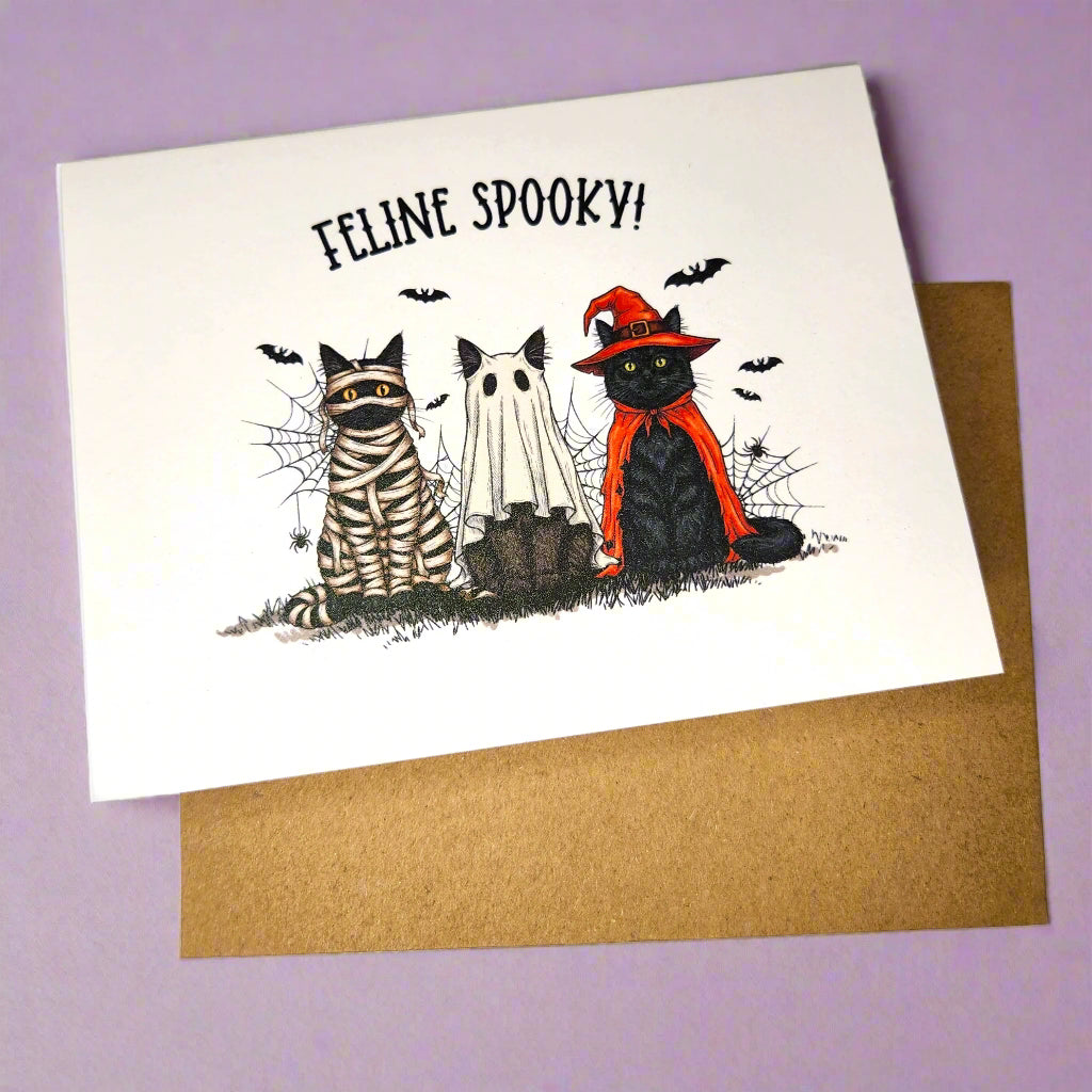 Halloween-themed card with cats dressed as a mummy, ghost, and witch on a purple background.