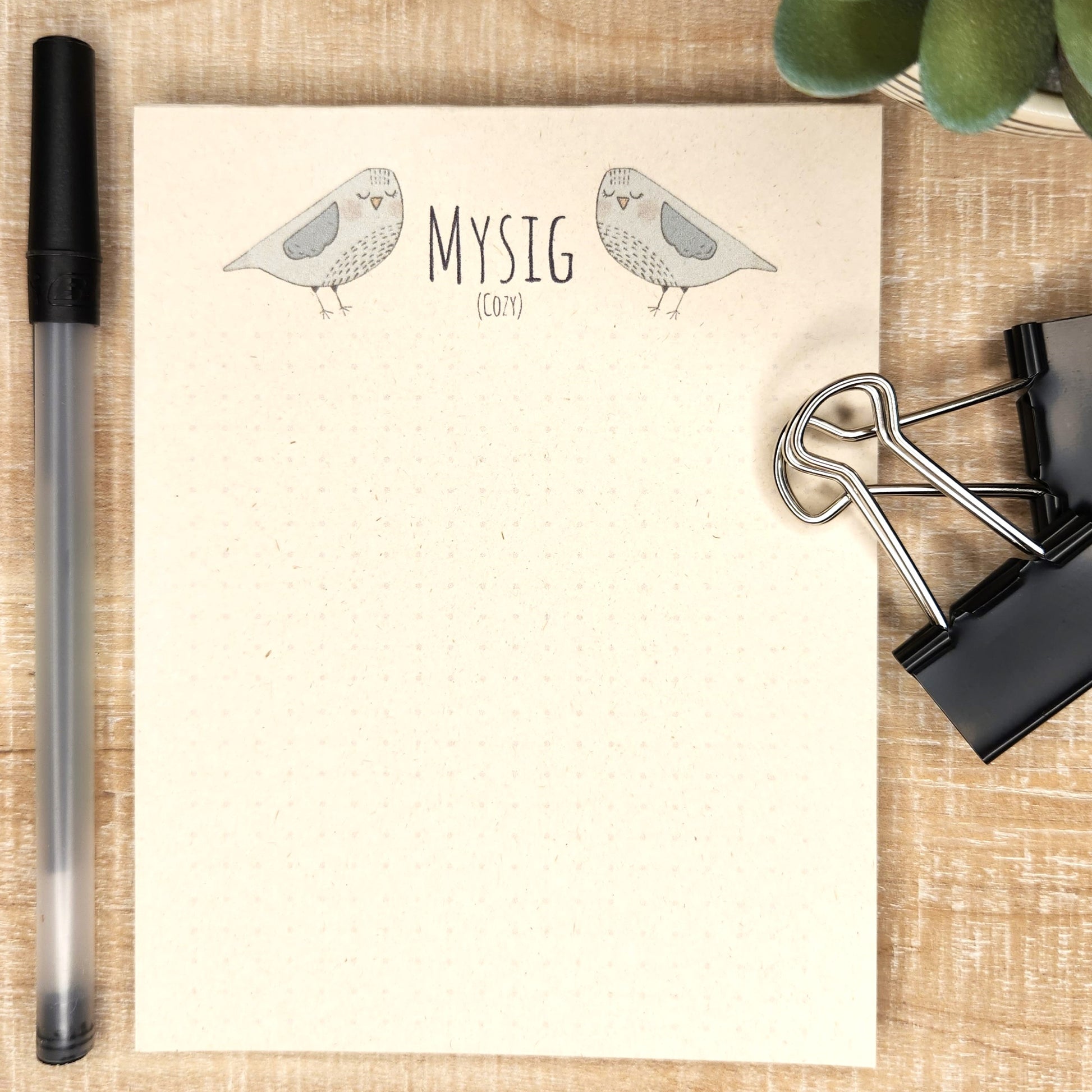 notepad on wooden background. blue/gray birds are on either side of the word 'Mysig' (cozy). A small dot grid fills out the bottom of the page.