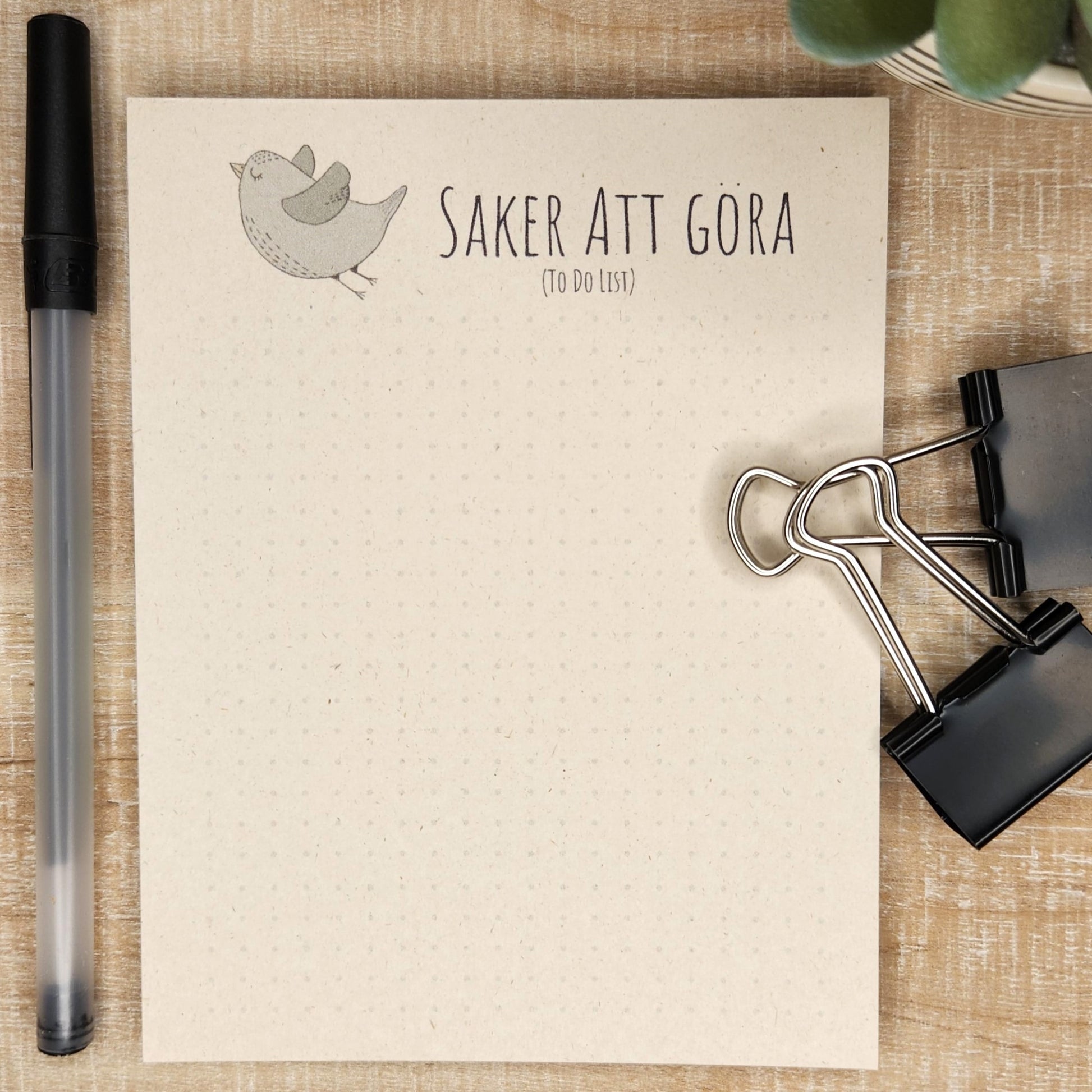 notepad on wooden background. green/gray bird flies off to the left of the words 'saker att gora' (to do list). A small dot grid fills out the bottom of the page.