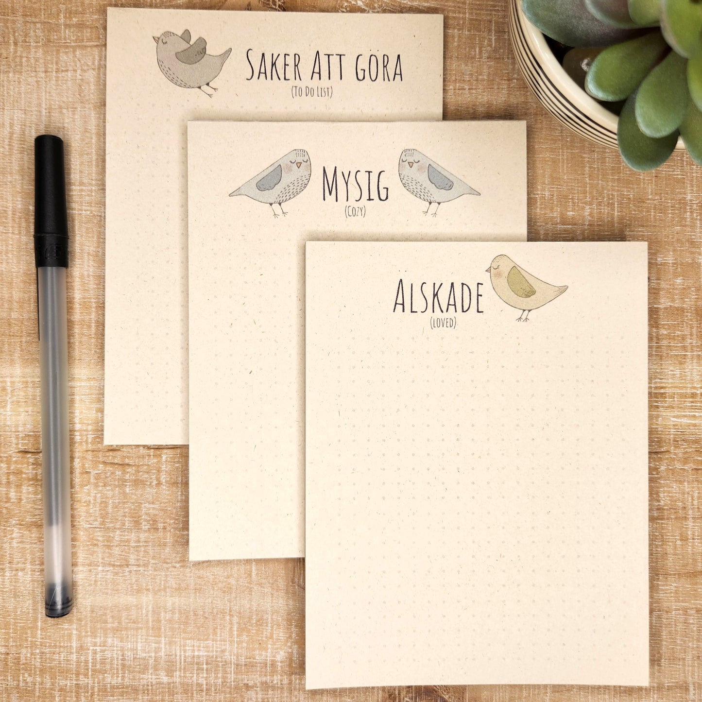 three Swedish inspired notepads lay on a wooden background. each designs show a Nordic inspired bird with Swedish word/words.