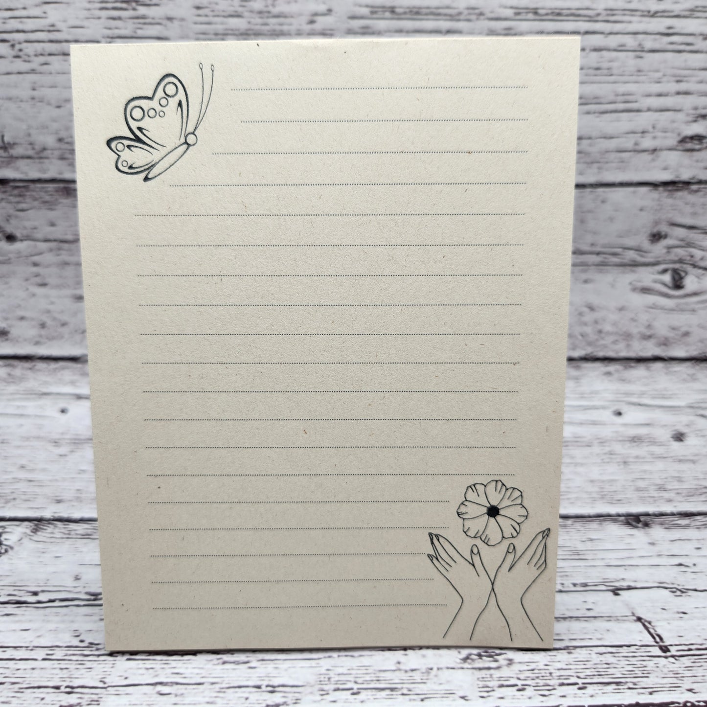 Soft beige sugarcane paper notepad on white barn board background.  Note pad is lined and has a butterfly accent on the upper left corner and hands with flowers on the bottom right corner.  all designs are line drawings. 