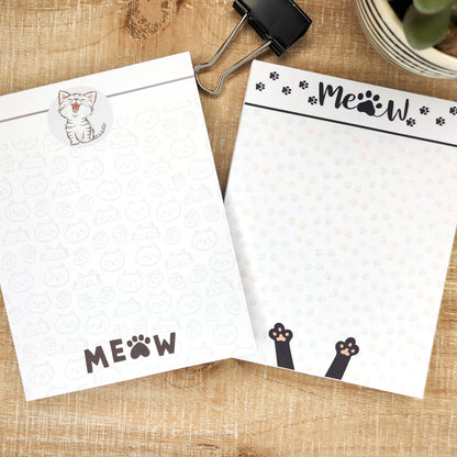 Set of two notepads are laying on a tan background. Notepads are white with 'meow' words and kitty and kitty paws.