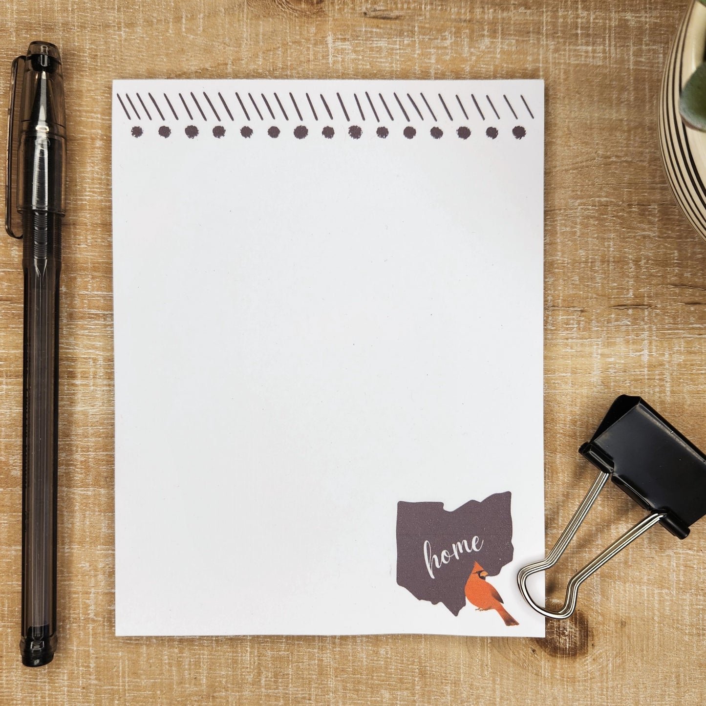 White notepad on a tan wood  background. Top of the page shows a festive banner of dots and lines. The solid black stat of Ohio is in the lower right hand side with the word home across the state. a red cardinal is on the right corner with the state.