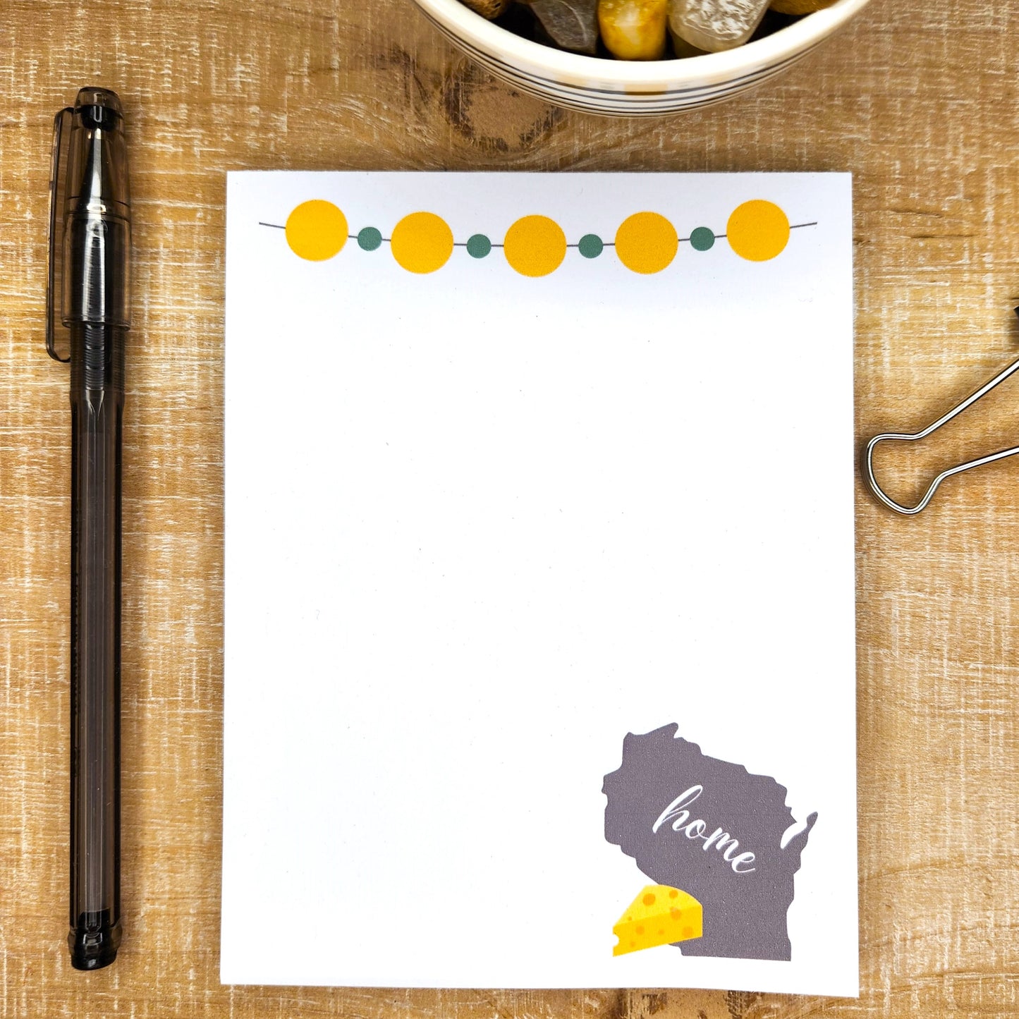 white notepad on wooden background. Notepad is topped with green and gold bead-looking banner. Bottom right side of pad shows the outline of the state of Wisconsin with the word home across it and a wedge of cheese on the side.