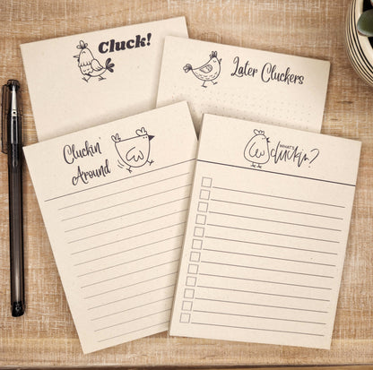 4 natural sugarcane notepads on a tan background. All four show a different cartoon chicken with a different saying. There is 'What's Cluckin?' 'Later Cluckers!' 'Cluckin' around' and 'Cluck!'