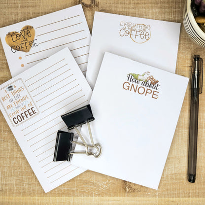 4 coffee related notepads are placed on a wooden background.  Each notepads has a different saying about coffee. 