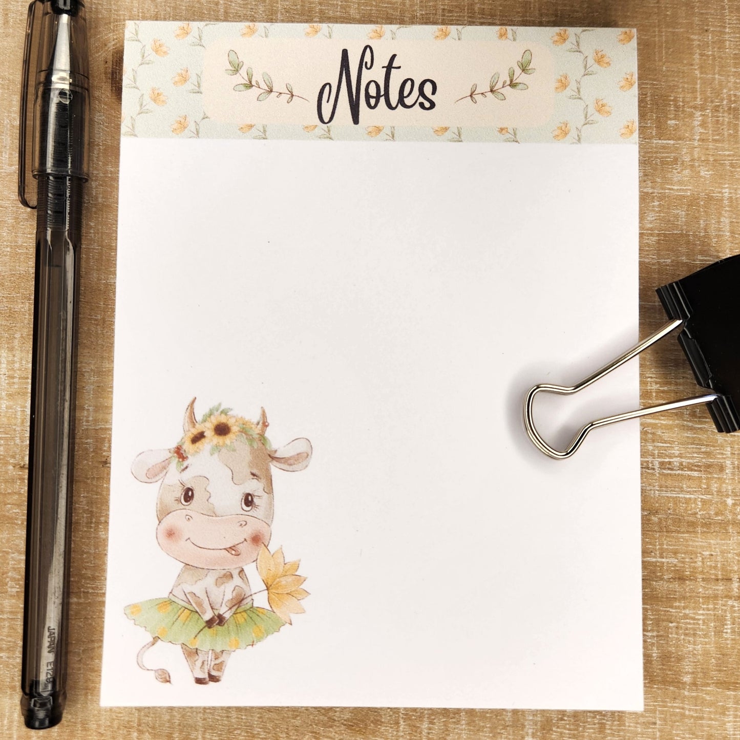 White notepad on wooden background. a floral border runs along the top inch of the page with the word Notes in the center. The lower right bottom corner show a cartoon cow in a tutu holding a flower.