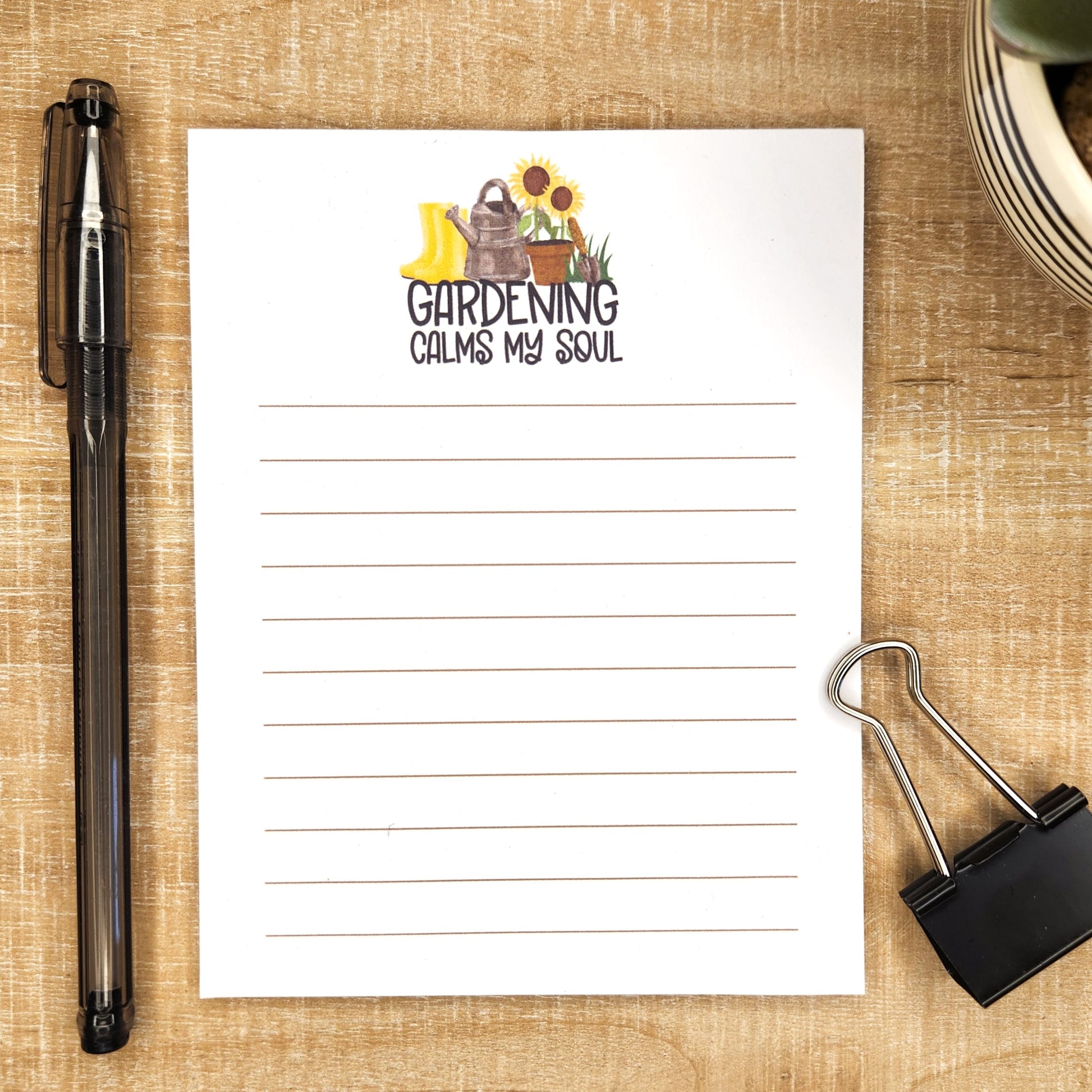 image of lined notepad on wooden background. words Gardening calms my soul with an image of boots, watering can, potted sunflower, and digger is above the words.