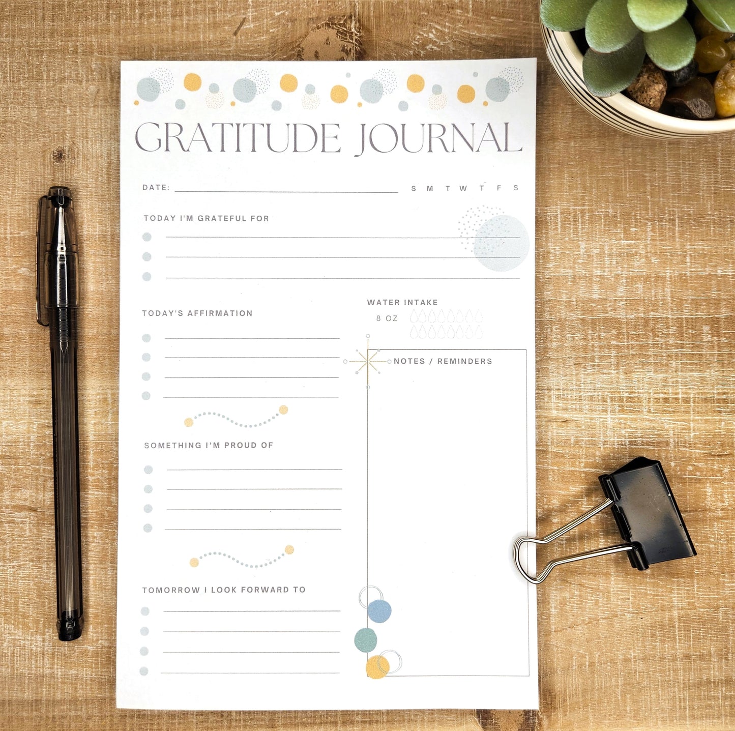 Beautiful half page gratitude journal printed on white paper and laying on a wooden background. Words on the page: gratitude Journal, date, today i'm grateful for, Today's affirmations, something I'm proud of, tomorrow I look forward to , water intake, notes/reminders.