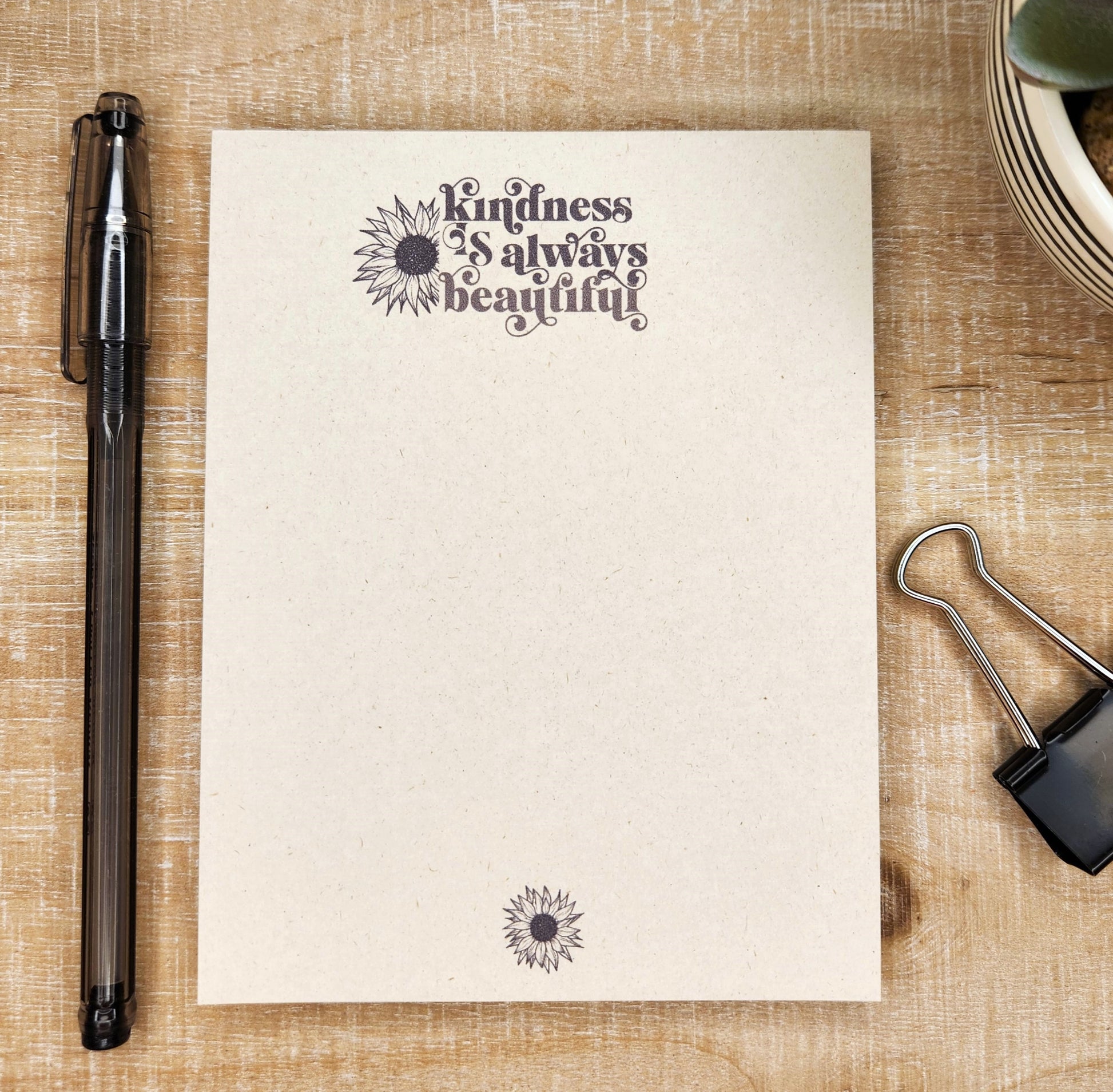 Tan sugarcane paper notepad on white barn board background. Notepad has the words 'Kindness is always beautiful'. a sunflower is to the left of the words and a small sunflower in on the bottom center of the page. all ink is black