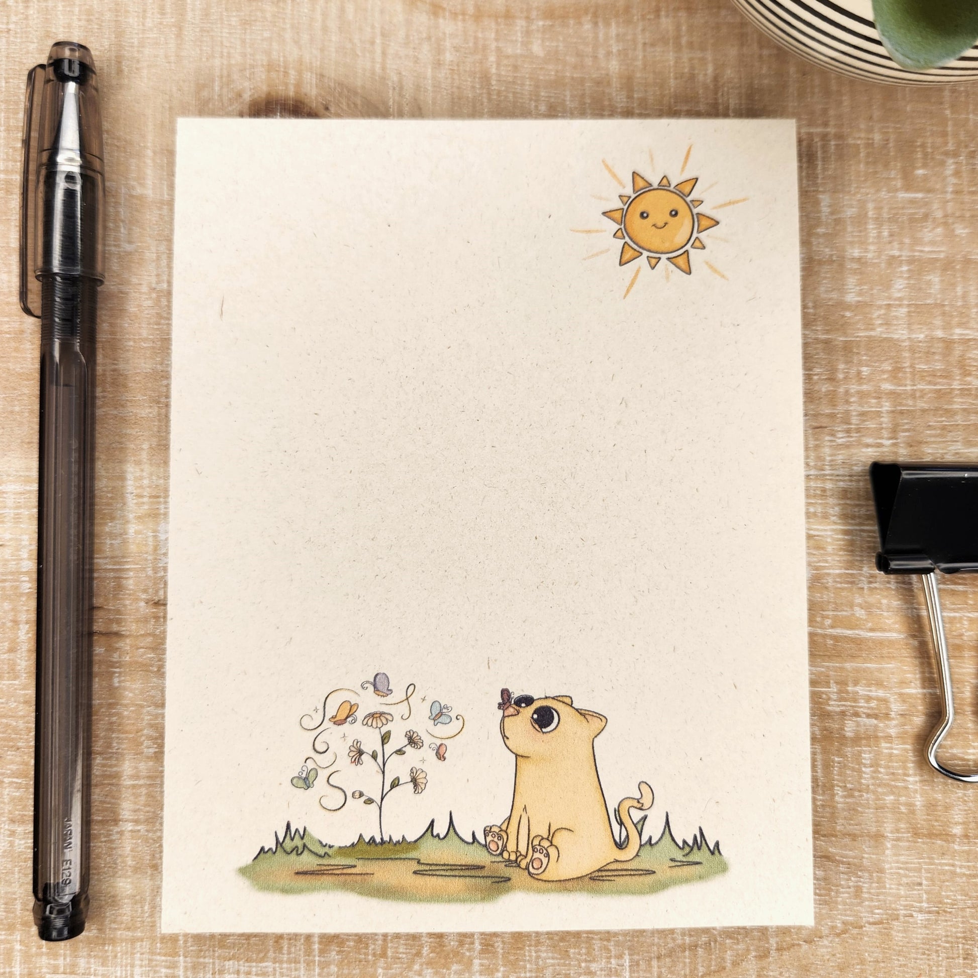 Natural sugarcane notepad on white background. Notepad depicts a sun in the upper right hand corner and sweet cartoon kitty with a butterfly on his nose is sitting on a patch of grass with a flower and butterflies.