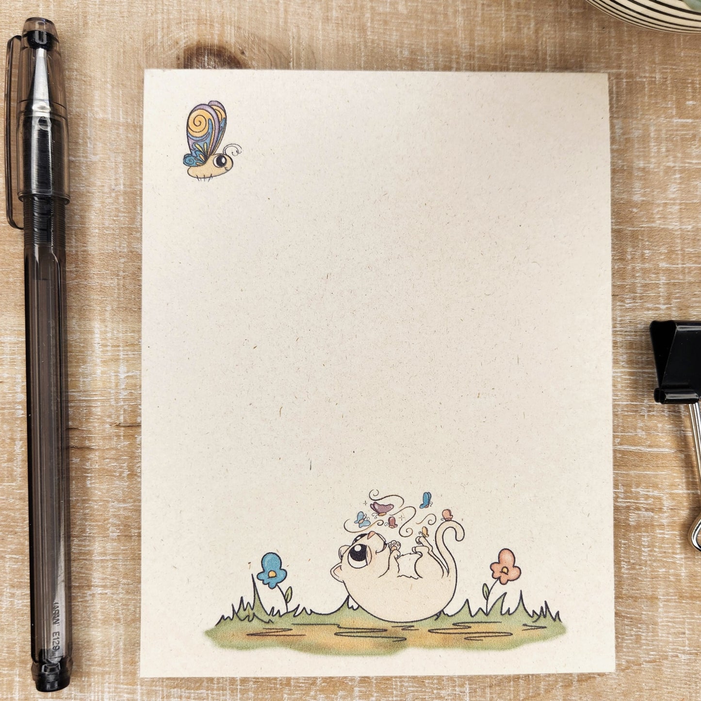 Natural sugarcane notepad on white background. Notepad depicts a butterfly in the upper left hand corner and sweet cartoon kitty laying on it's back with a butterfly on flying over it. Kitty is on a patch of grass with a flower and butterflies.