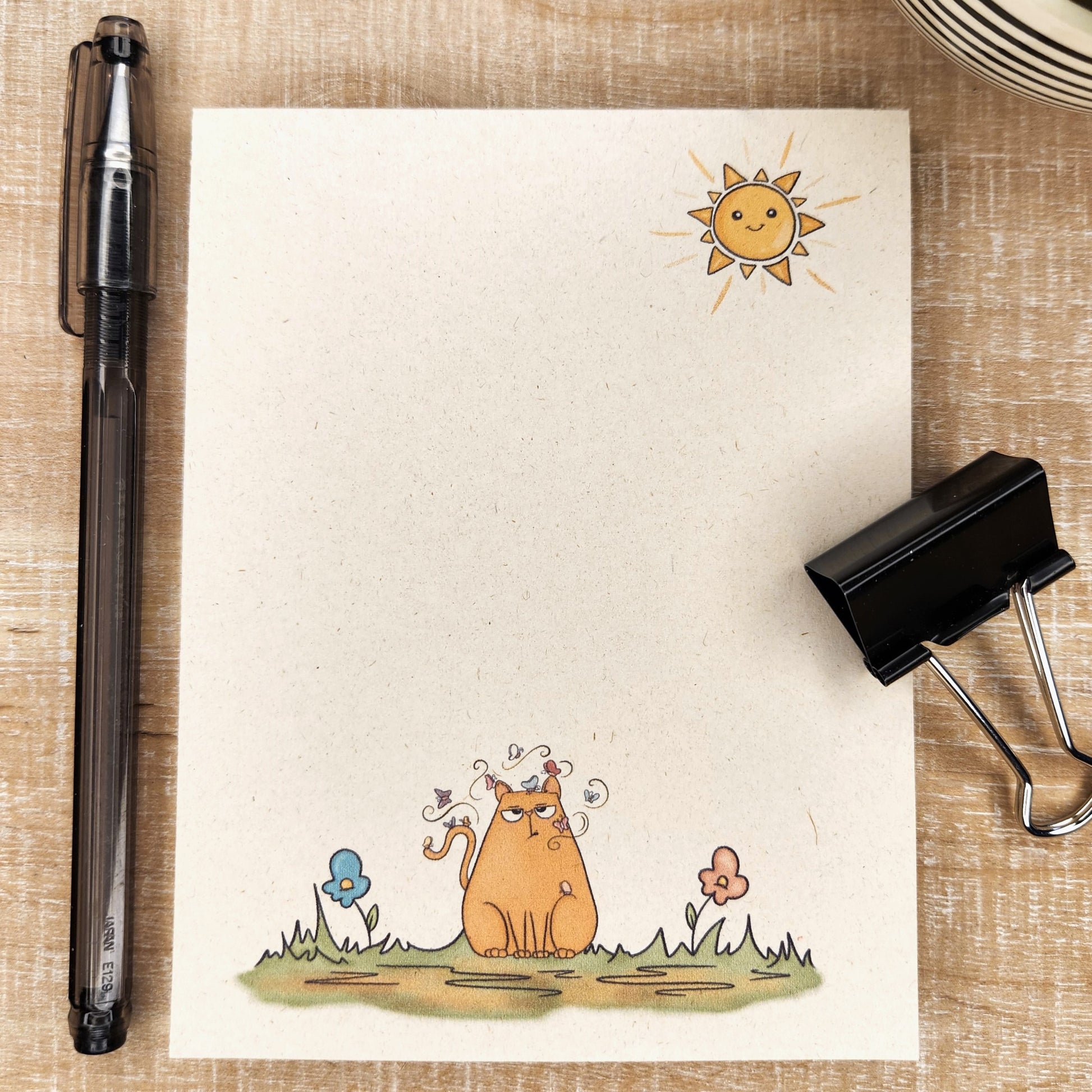 Natural sugarcane notepad on white background. Notepad depicts a sun in the upper right hand corner and an unhappy cartoon kitty with a butterfly on his head is sitting on a patch of grass with butterflies circling the kitty's head.