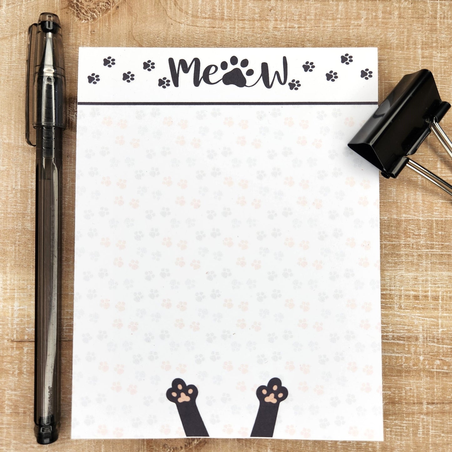 white notepad on tan background. Word Meow with kitty prints are across the top of the notepad with a black line accent. Faint kitty prints in pinks and greys fill the page with black cat paws are in the center bottom of the page.