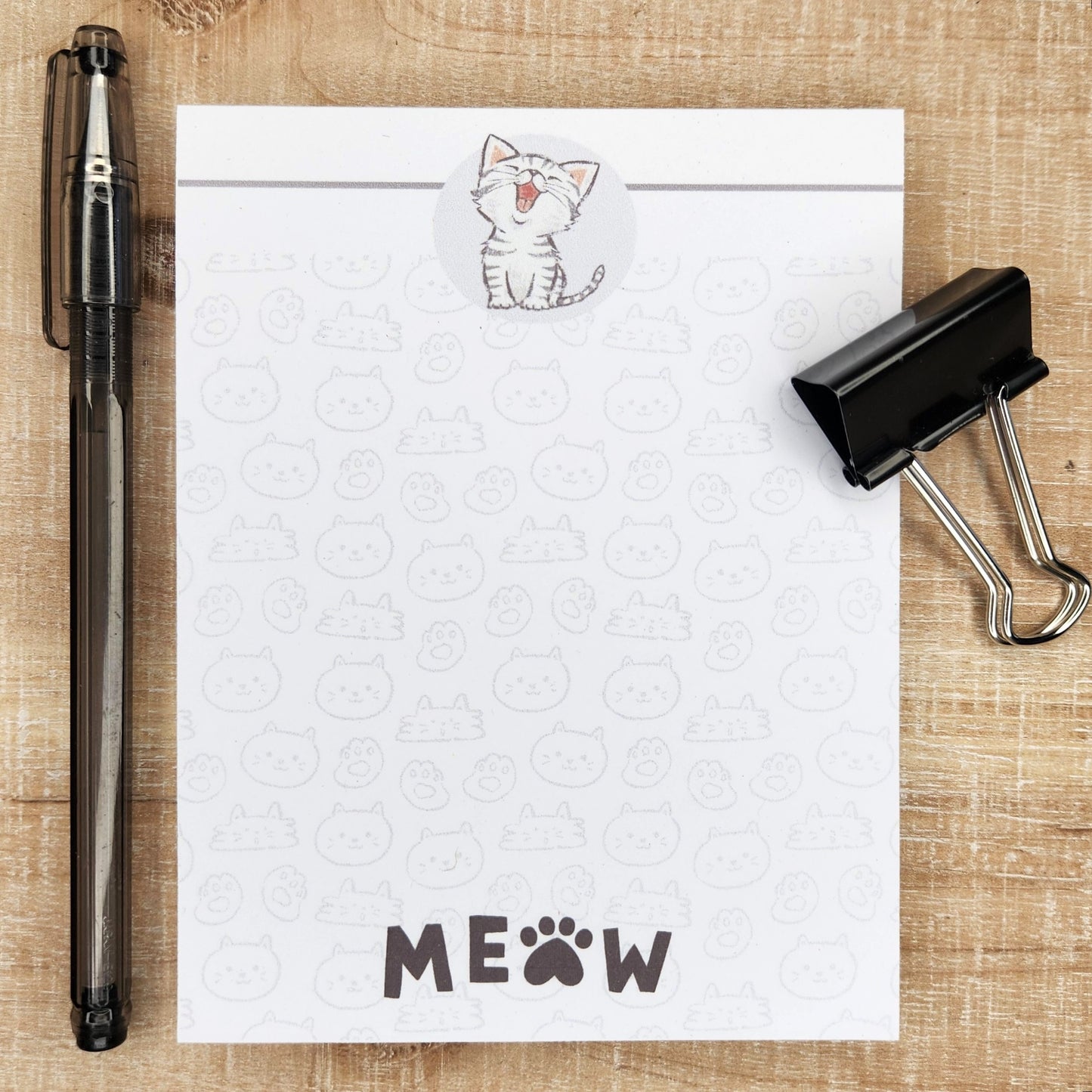 White notepad on a tan background. Kitten with mouth open graces the top center of the page. Black line accents the top as well. Faint cat faces are throughout the page and the word MEOW in on the center bottom of the page.