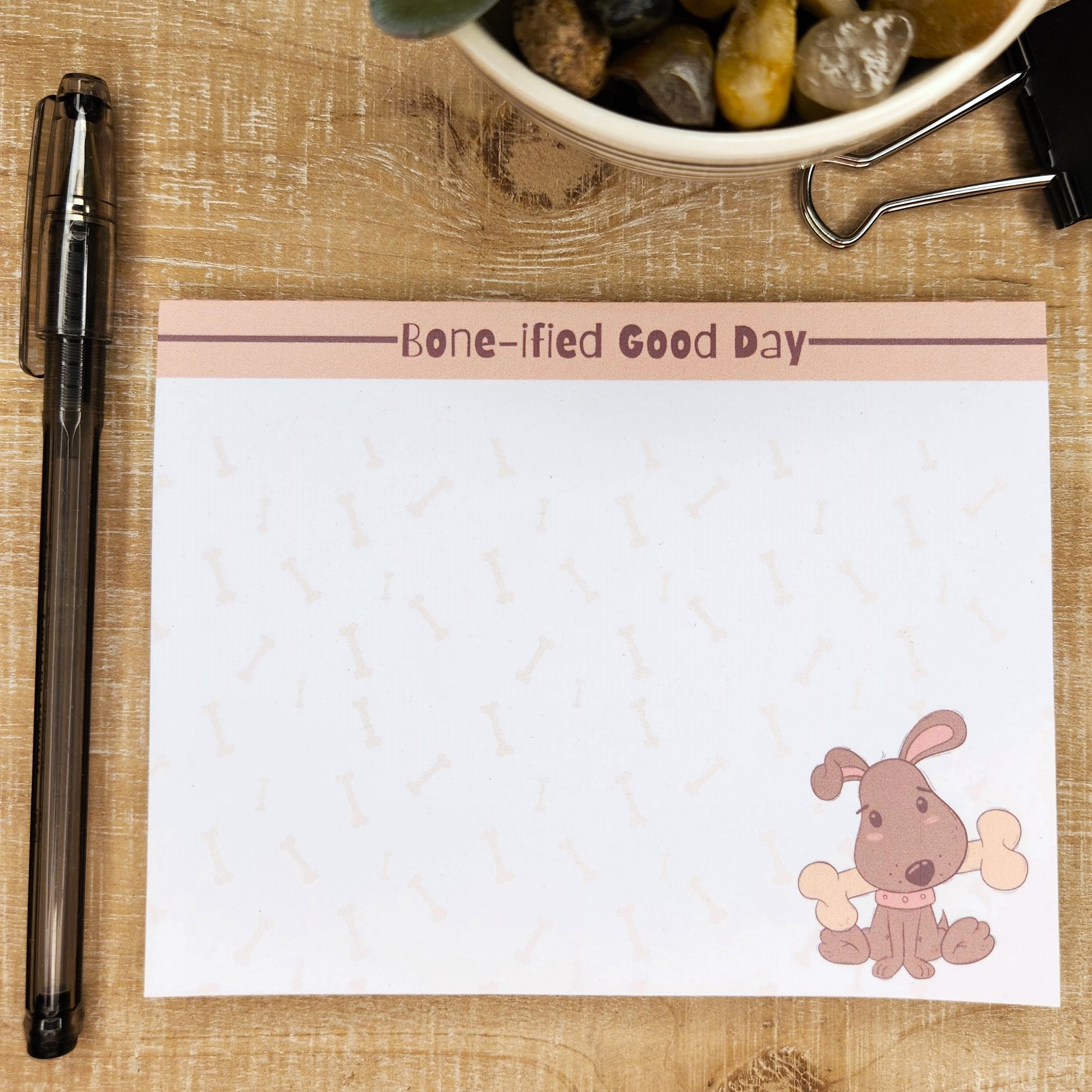 White notepad on blue background. Top of page has blush accent bar with burgundy line. The words on the center top of the page are ""Bone-ified Good Day". Puppy with bone on the bottom right side of the page. Faint dog bones are though out the page.