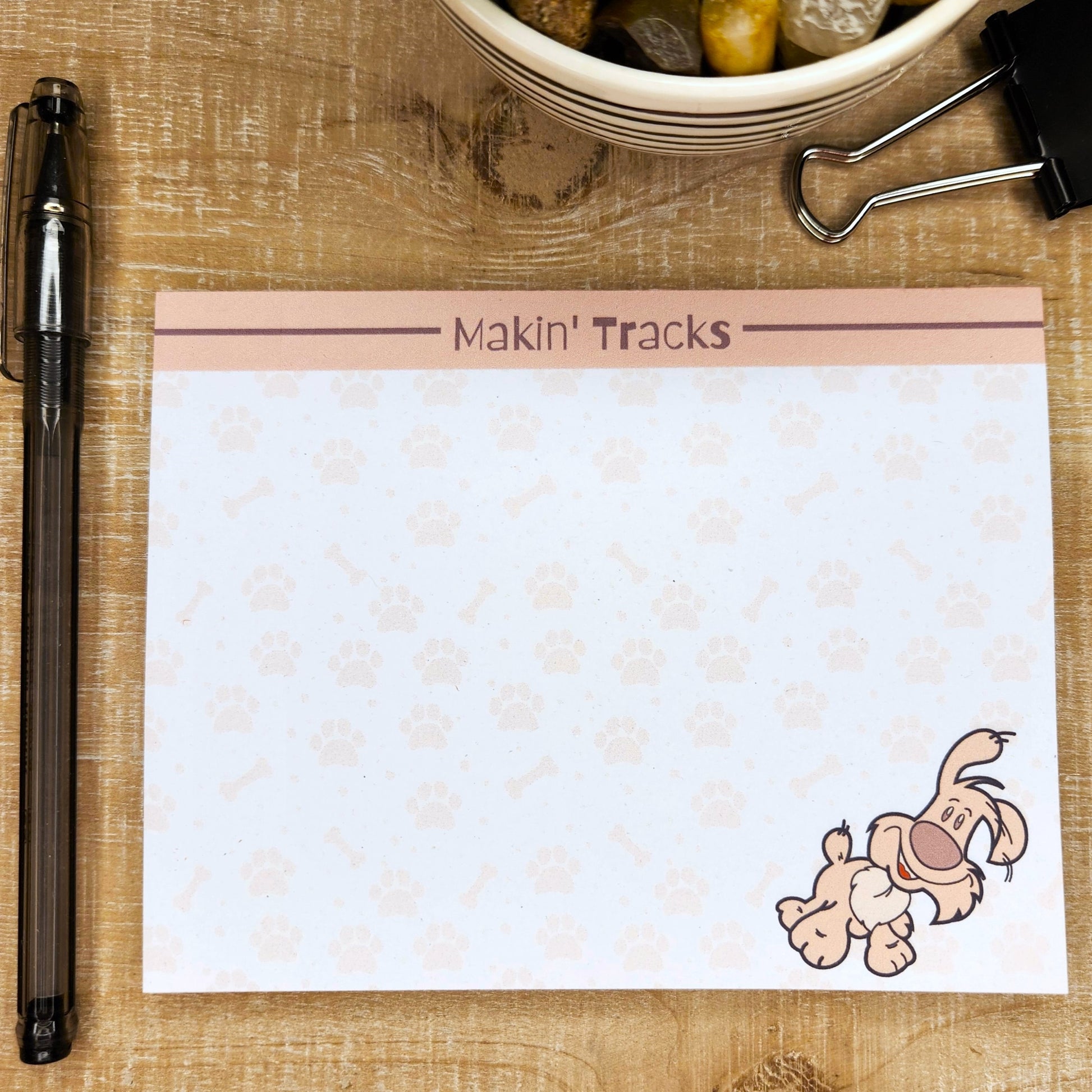 White notepad on tan background. Top of page has blush accent bar with burgundy line. The words on the center top of the page are ""Makin' Tracks". Silly puppy on the bottom right side of the page. Faint dog tracks and bones are though out the page.