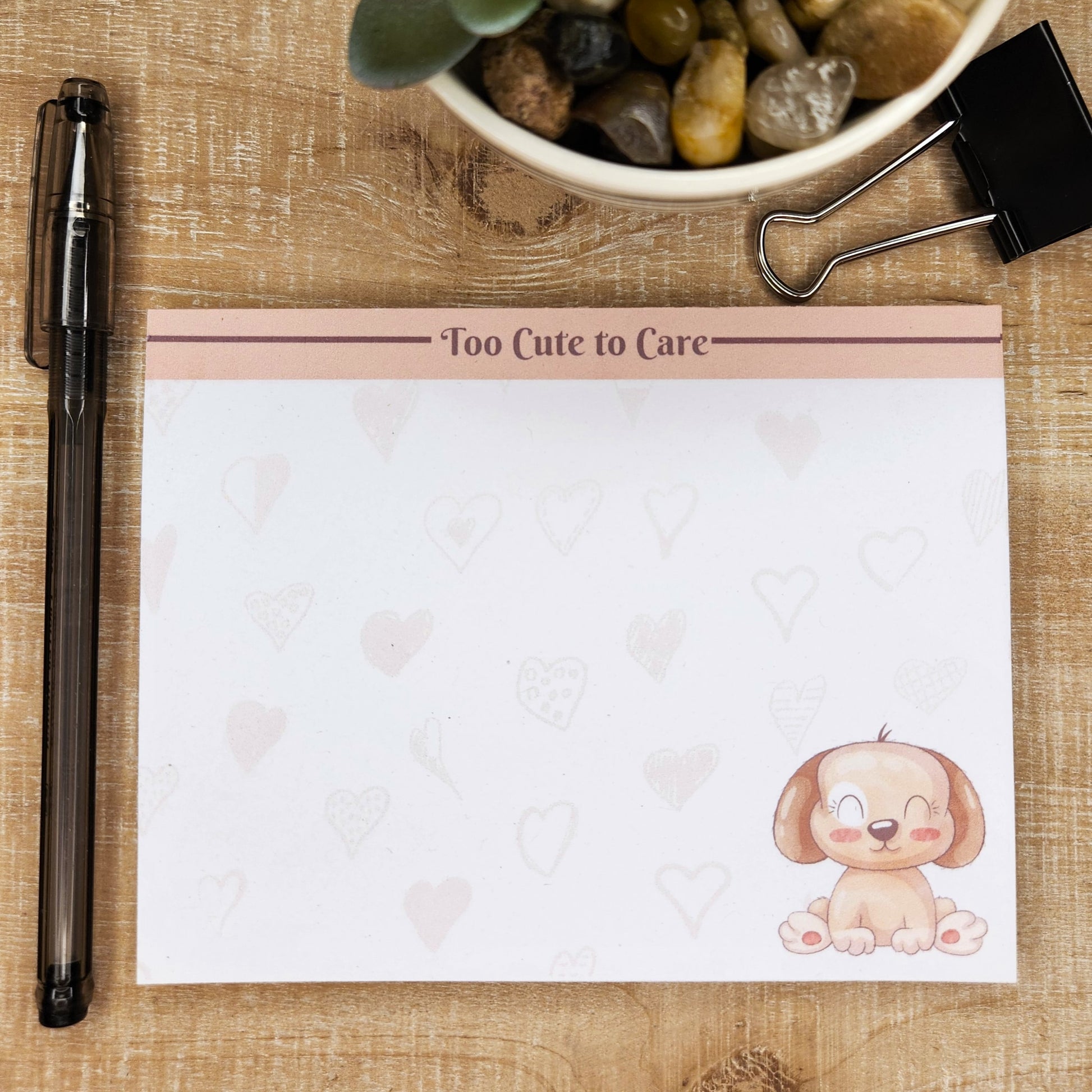 White notepad on tan background. Top of page has blush accent bar with burgundy line. The words on the center top of the page are ""Too cute to care". Sitting puppy on the bottom right side of the page. Faint hearts are though out the page.