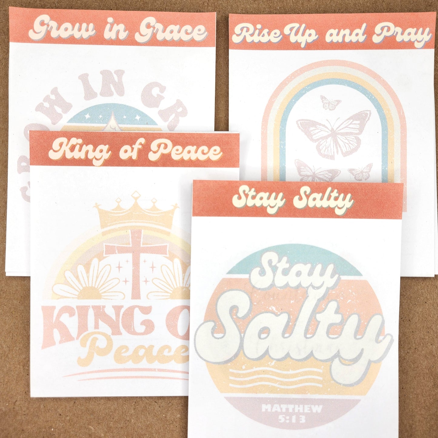 4 white notepads on a tan background. Each notepad has a banner in orange on the top of the page. Stay Salty, Rise up and pray, King of peace, Grow in Grace all in a vintage 70 vibe.