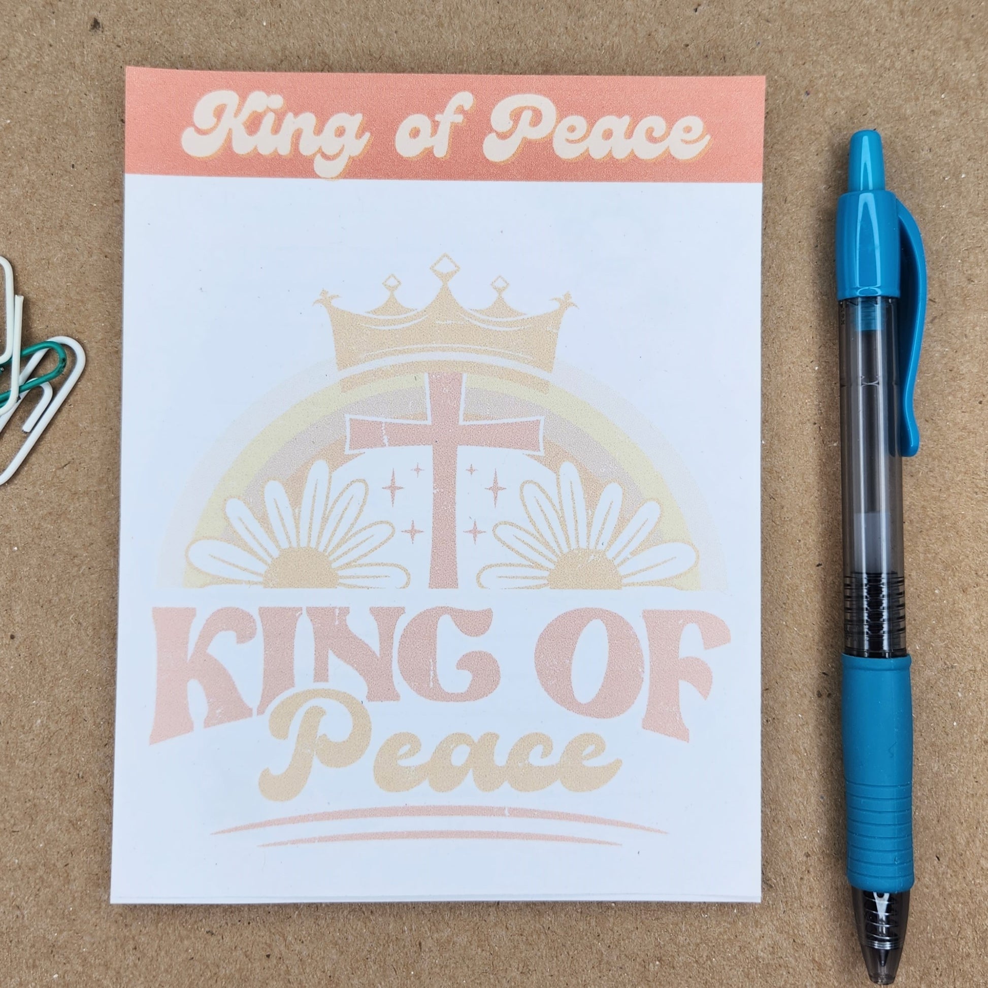 White notepad on a tan background. Words king of peace in a retro style. Center of notepad has a cross and flowers with the words king of peace.