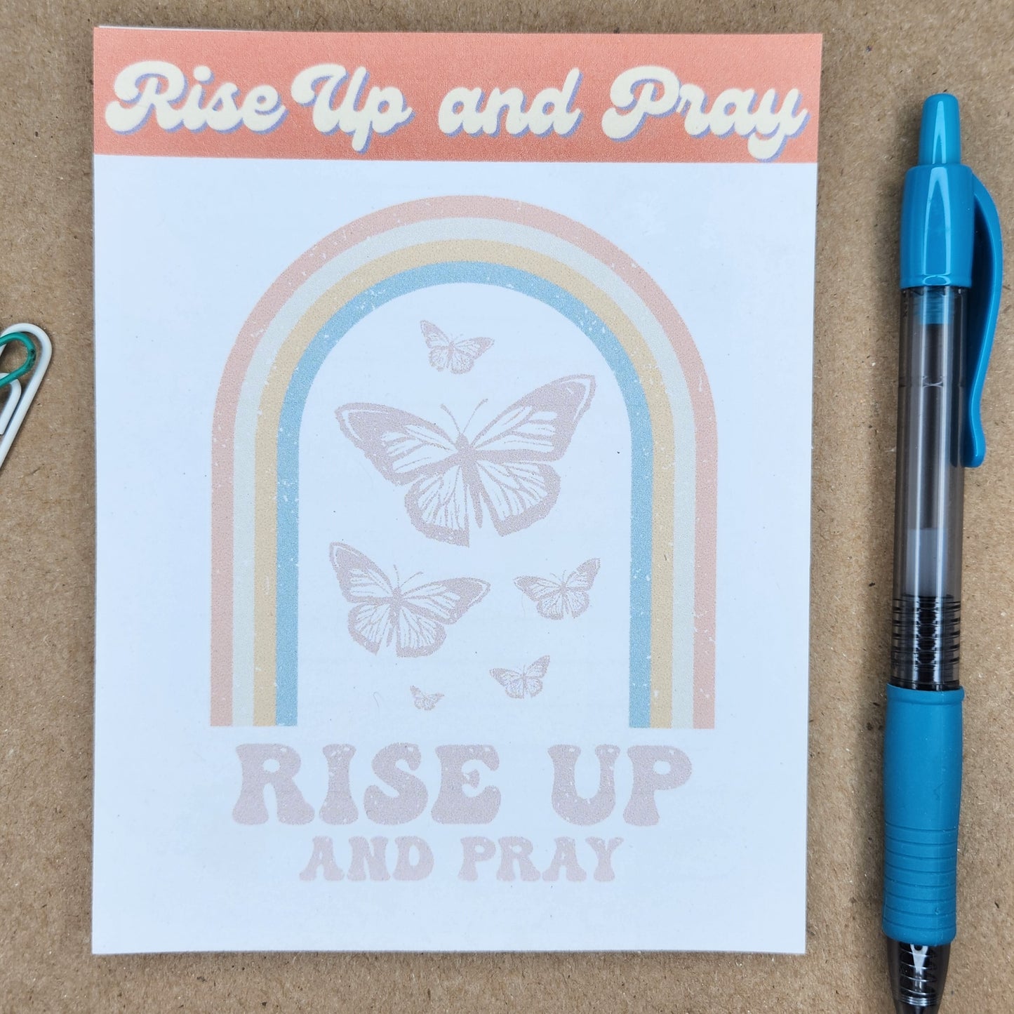 White notepad on a tan background. Words Rise up and pray in a retro style. Center of notepad has a rainbow and butterflies with the words rise up and pray.