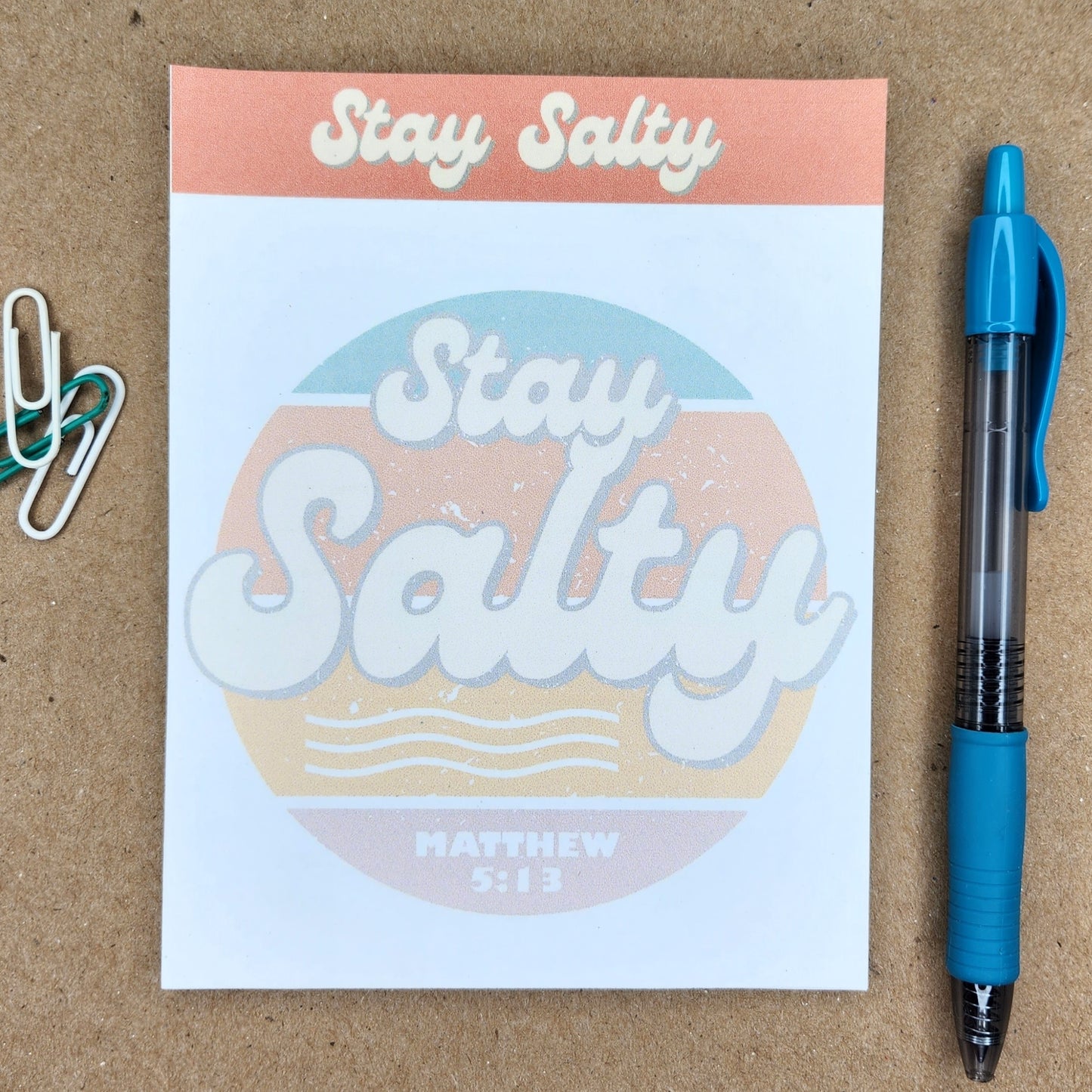 White notepad on a tan background. Words Stay Salty in a retro style. Center of notepad has a circle and the words Stay Salty