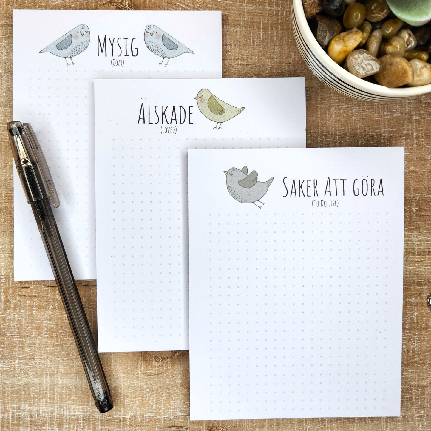 three Swedish inspired notepads lay on a wooden background.  each designs show a Nordic inspired bird with Swedish word/words.