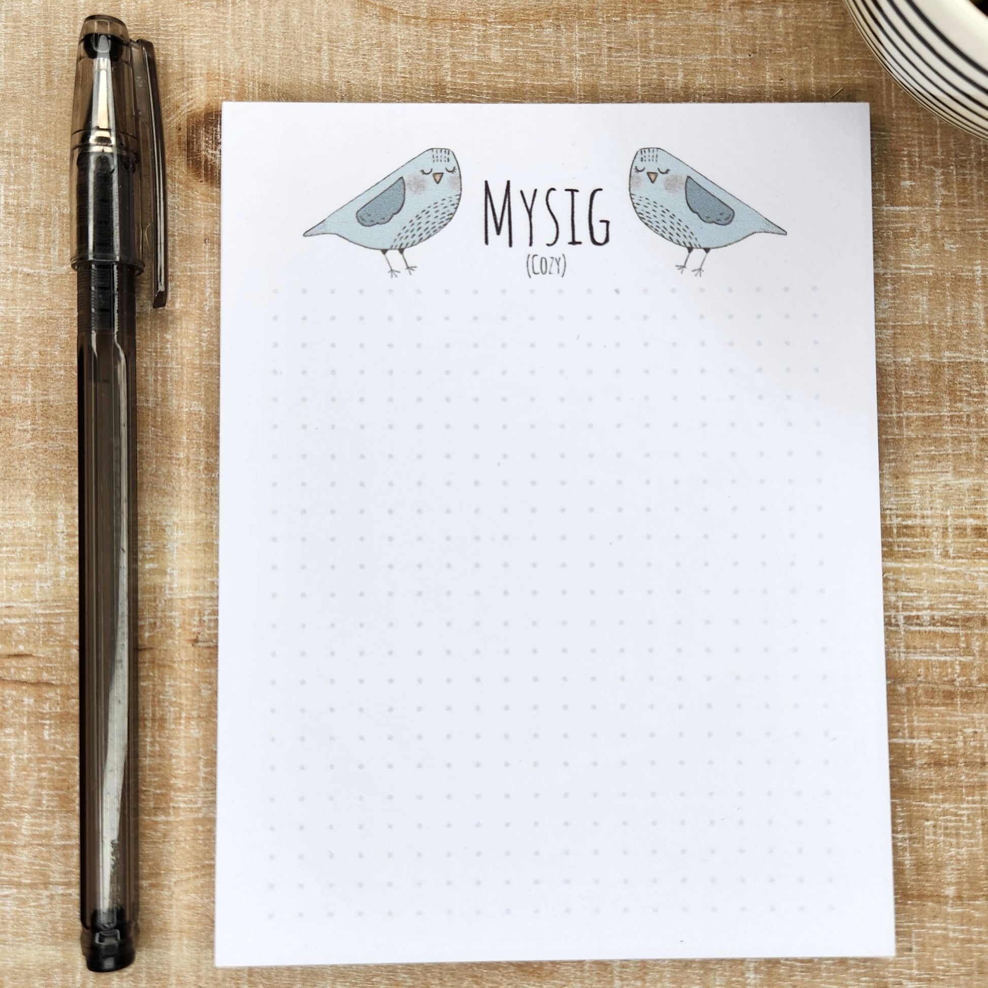 notepad on wooden background. blue/gray birds are on either side  of the word 'Mysig' (cozy). A small dot grid fills out the bottom of the page.