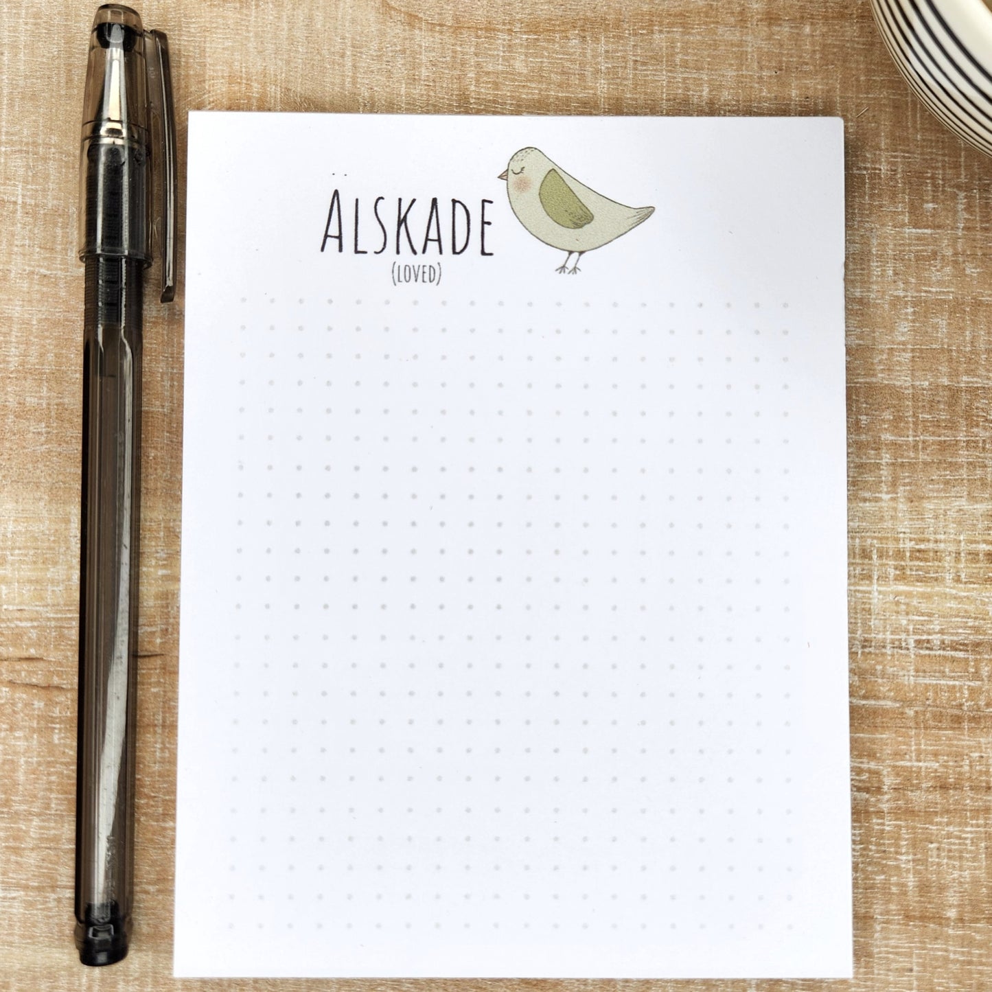 notepad on wooden background. green/gray bird sits to the right  side of the word 'Alskade' (loved). A small dot grid fills out the bottom of the page.