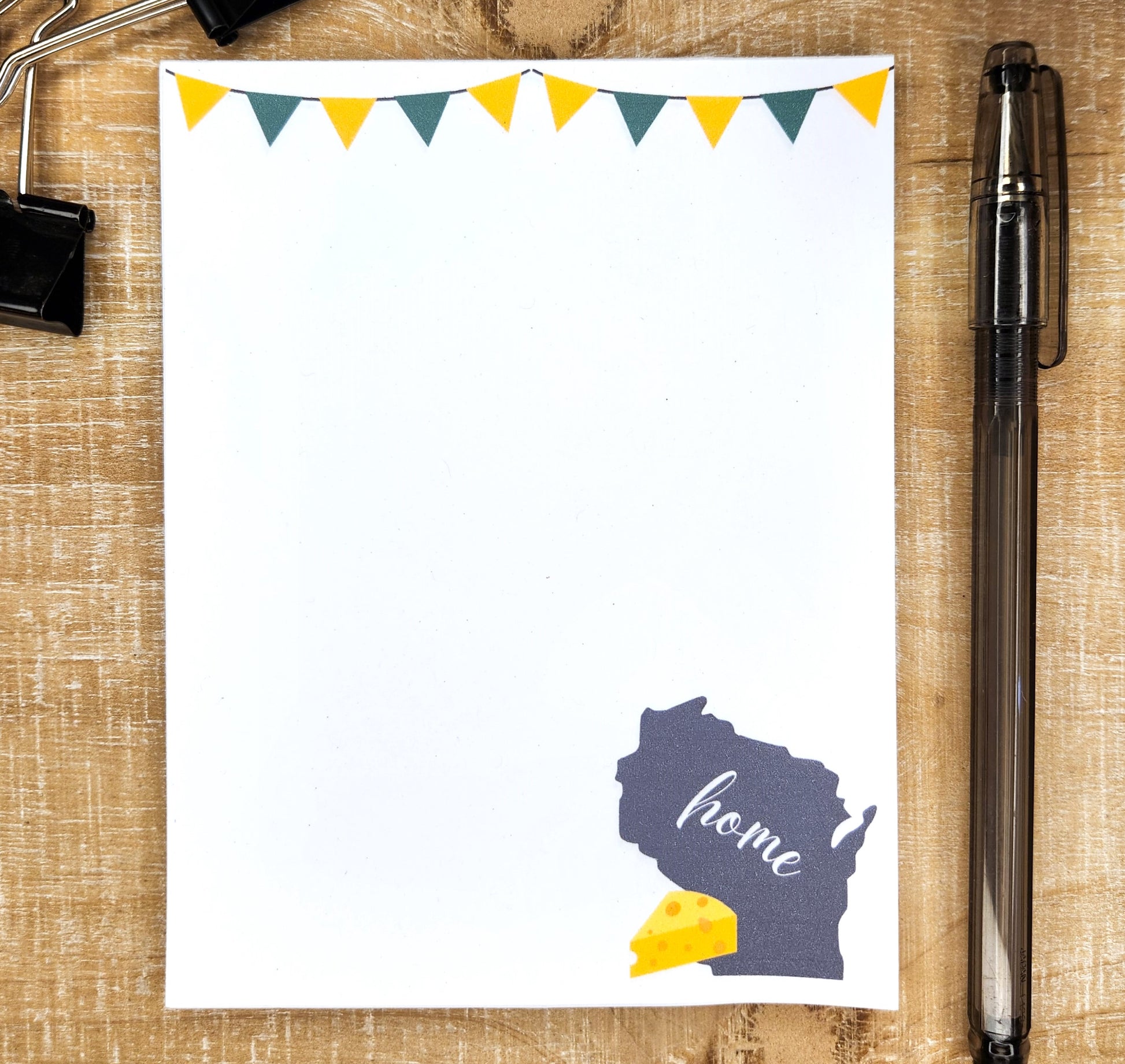 white notepad on tan background. Notepad is topped with green and gold flag banner. Bottom right side of pad shows the outline of the state of Wisconsin with the word home across it and a wedge of cheese on the side.